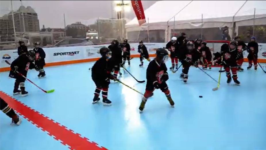 Children in hockey gear playing a scrimmage on the synthetic ice surface, sticks down and a puck in play, with boards and netting around the rink.