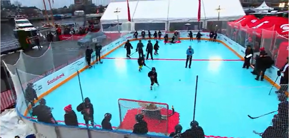 The full rink set up outdoors — synthetic ice surface enclosed by dasher boards and netting, with skaters on it.